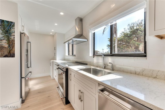 kitchen featuring white cabinetry, appliances with stainless steel finishes, wall chimney range hood, light stone countertops, and sink