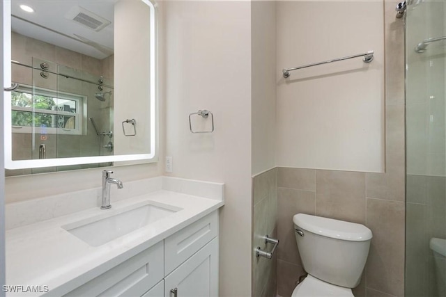 bathroom with visible vents, toilet, a tile shower, vanity, and tile walls