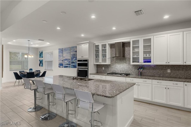 kitchen with appliances with stainless steel finishes, white cabinetry, an island with sink, backsplash, and wall chimney exhaust hood