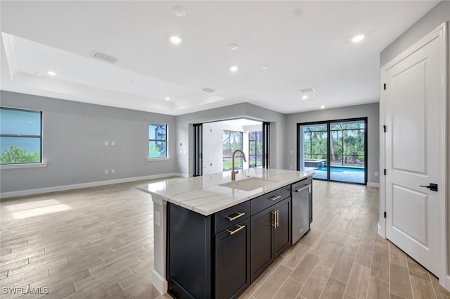 kitchen featuring an island with sink, sink, a raised ceiling, light stone counters, and stainless steel dishwasher