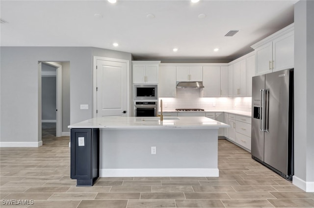 kitchen with white cabinetry, stainless steel appliances, a center island with sink, and light stone countertops