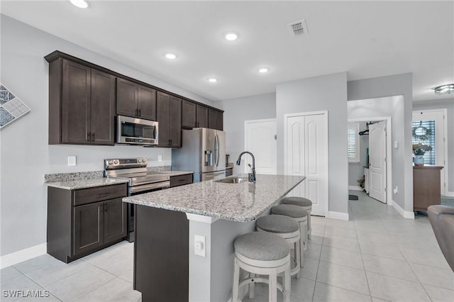 kitchen with light stone countertops, dark brown cabinetry, appliances with stainless steel finishes, an island with sink, and sink