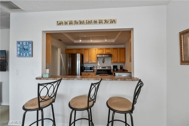 kitchen featuring kitchen peninsula, a breakfast bar area, appliances with stainless steel finishes, a raised ceiling, and dark stone countertops