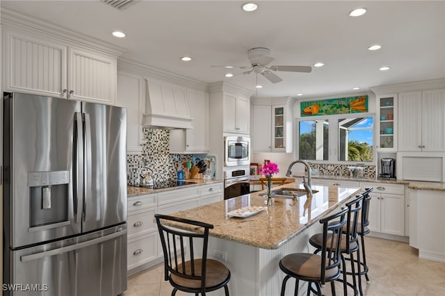 kitchen with custom exhaust hood, a center island with sink, appliances with stainless steel finishes, light stone countertops, and sink