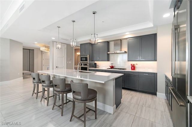 kitchen with appliances with stainless steel finishes, a raised ceiling, a center island with sink, and wall chimney range hood