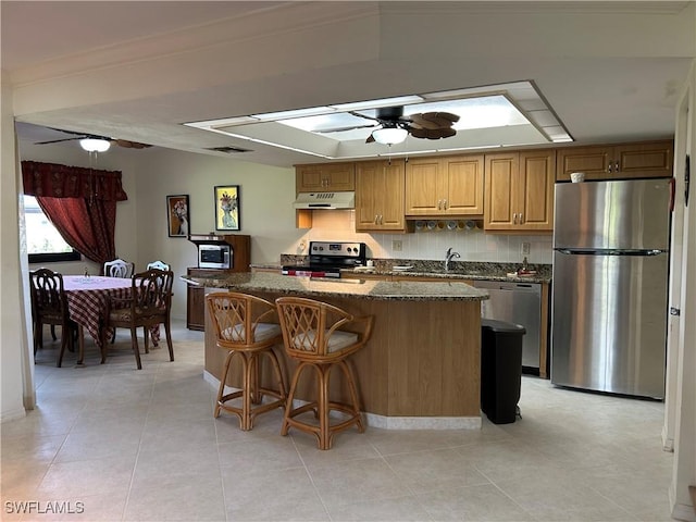 kitchen featuring a kitchen bar, dark stone counters, ceiling fan, stainless steel appliances, and backsplash