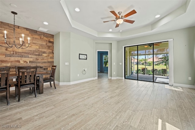 unfurnished living room with a raised ceiling, ceiling fan with notable chandelier, light hardwood / wood-style flooring, and wood walls