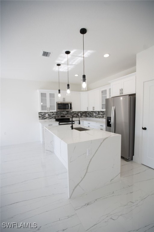 kitchen with white cabinetry, appliances with stainless steel finishes, pendant lighting, light stone countertops, and a kitchen island with sink