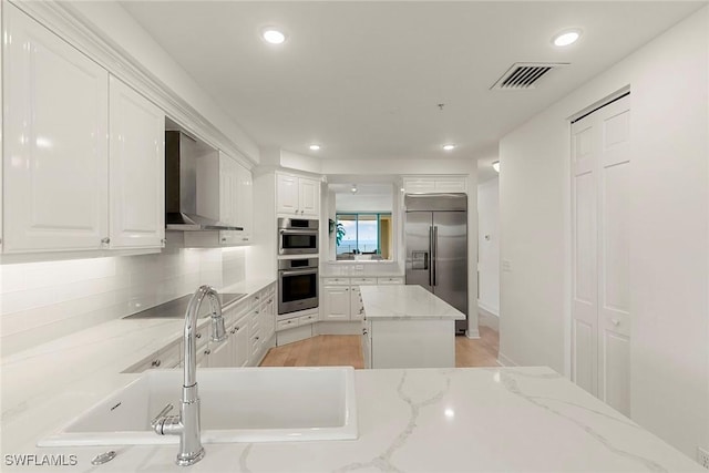 kitchen featuring wall chimney range hood, white cabinetry, light stone counters, and appliances with stainless steel finishes