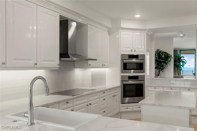 kitchen featuring light stone countertops, stainless steel double oven, white cabinetry, wall chimney range hood, and black electric stovetop