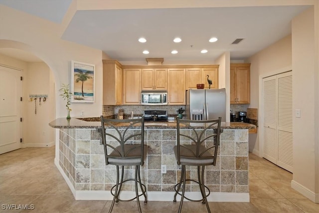 kitchen featuring stainless steel appliances, decorative backsplash, light brown cabinetry, light tile patterned flooring, and a breakfast bar area