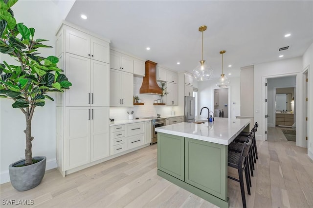 kitchen featuring a center island with sink, white cabinetry, hanging light fixtures, appliances with stainless steel finishes, and custom range hood