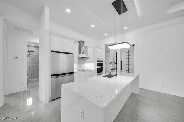 kitchen featuring decorative light fixtures, white cabinetry, a kitchen island with sink, appliances with stainless steel finishes, and wall chimney exhaust hood