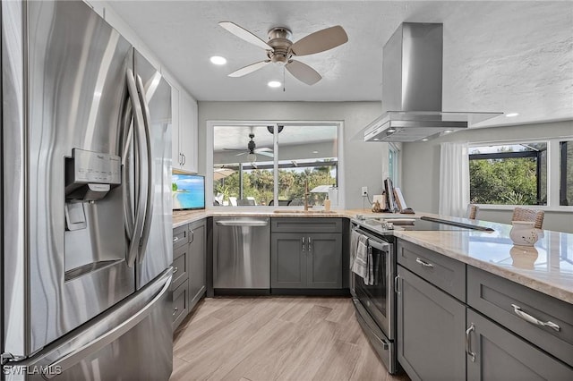 kitchen with island exhaust hood, appliances with stainless steel finishes, gray cabinetry, light wood-type flooring, and light stone counters