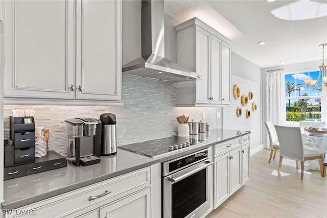 kitchen featuring wall chimney range hood, hanging light fixtures, stainless steel oven, light hardwood / wood-style flooring, and black electric cooktop