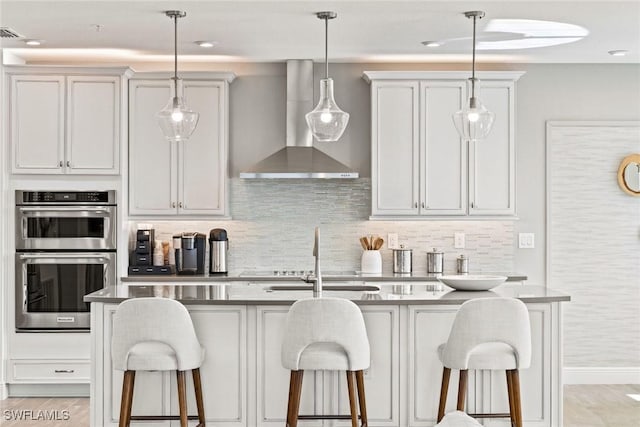 kitchen with white cabinetry, wall chimney range hood, double oven, a kitchen breakfast bar, and a kitchen island with sink