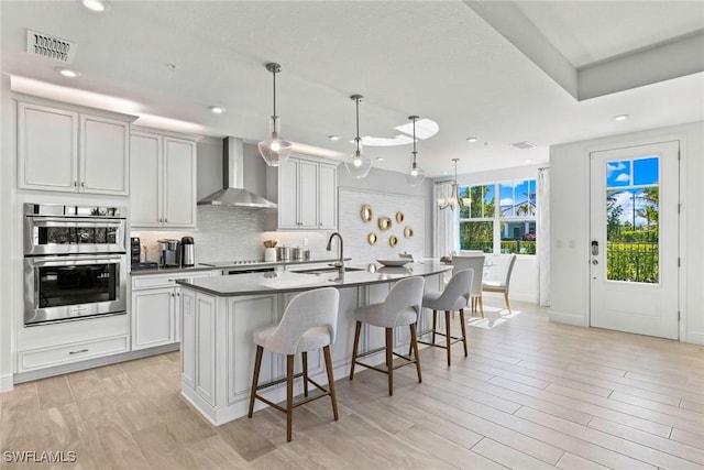 kitchen with a center island with sink, wall chimney range hood, pendant lighting, white cabinets, and sink