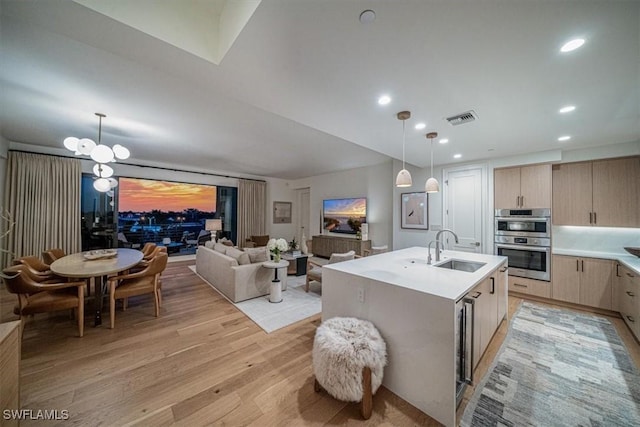 kitchen featuring an island with sink, decorative light fixtures, light brown cabinetry, double oven, and sink