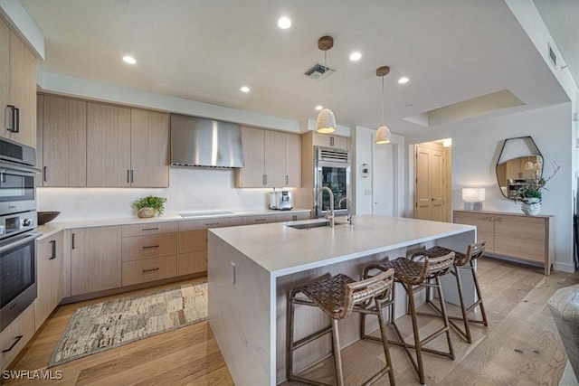 kitchen featuring light hardwood / wood-style floors, a kitchen island with sink, light brown cabinetry, wall chimney range hood, and sink