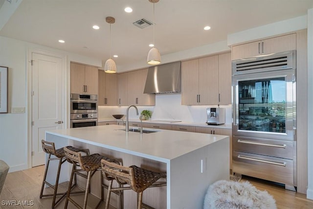 kitchen with decorative light fixtures, wall chimney range hood, sink, light wood-type flooring, and a kitchen island with sink