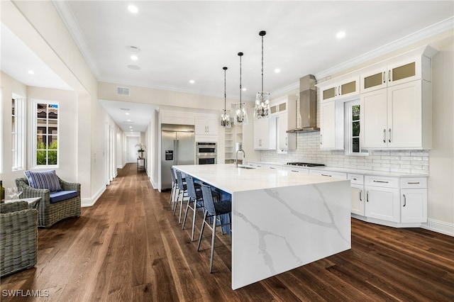 kitchen featuring white cabinetry, wall chimney range hood, sink, stainless steel appliances, and a large island with sink