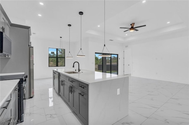 kitchen featuring sink, hanging light fixtures, gray cabinets, a tray ceiling, and a center island with sink