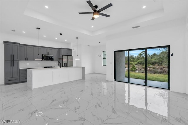 kitchen featuring decorative light fixtures, stainless steel appliances, a raised ceiling, a kitchen island with sink, and gray cabinetry