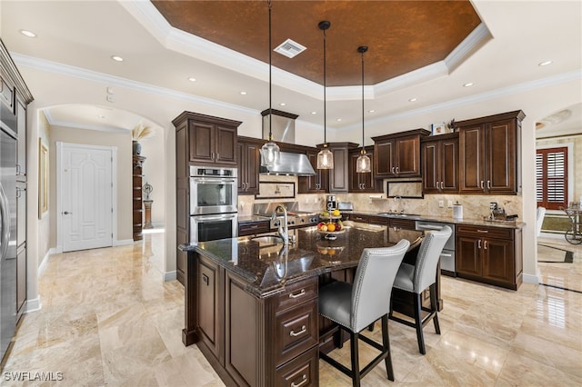 kitchen featuring pendant lighting, an island with sink, dark stone countertops, dark brown cabinetry, and a tray ceiling