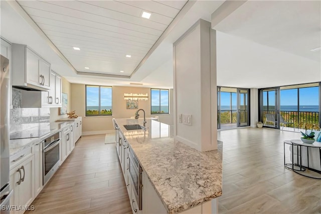 kitchen featuring a kitchen island with sink, white cabinetry, light stone countertops, a raised ceiling, and stainless steel oven