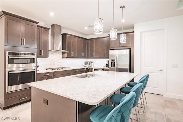 kitchen featuring wall chimney exhaust hood, appliances with stainless steel finishes, dark brown cabinetry, a sink, and backsplash