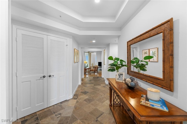 hallway featuring stone finish flooring, a tray ceiling, and baseboards