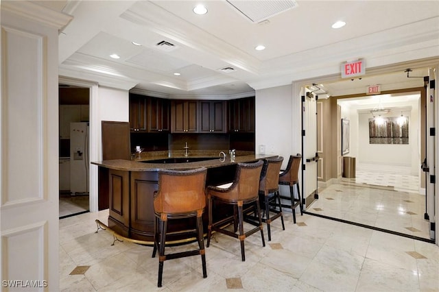bar with kitchen peninsula, dark brown cabinetry, crown molding, white refrigerator with ice dispenser, and a kitchen bar