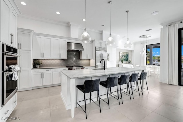 kitchen with white cabinets, decorative light fixtures, stainless steel appliances, an island with sink, and ventilation hood