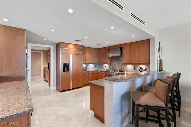 kitchen with a breakfast bar, dark stone counters, paneled built in refrigerator, kitchen peninsula, and wall chimney range hood