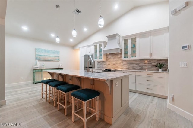 kitchen featuring premium range hood, white cabinetry, hanging light fixtures, light stone countertops, and a center island with sink