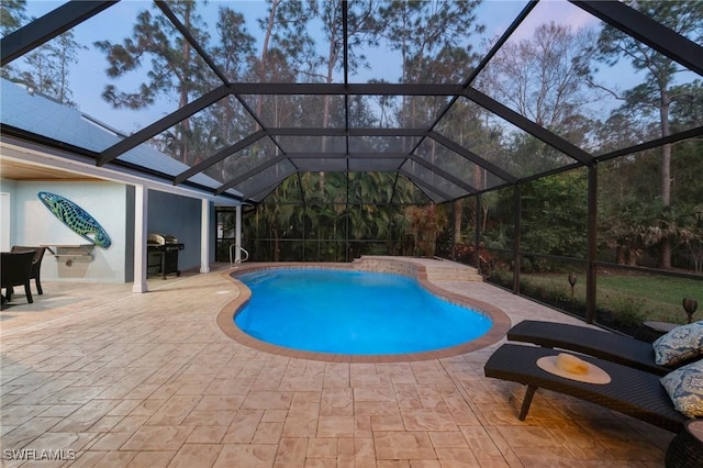 pool at dusk featuring area for grilling, a patio area, and glass enclosure