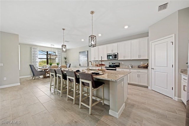 kitchen featuring a center island with sink, stainless steel appliances, white cabinets, hanging light fixtures, and light stone counters