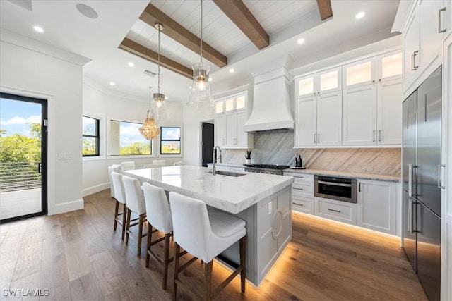 kitchen with custom exhaust hood, sink, stainless steel built in fridge, white cabinetry, and an island with sink