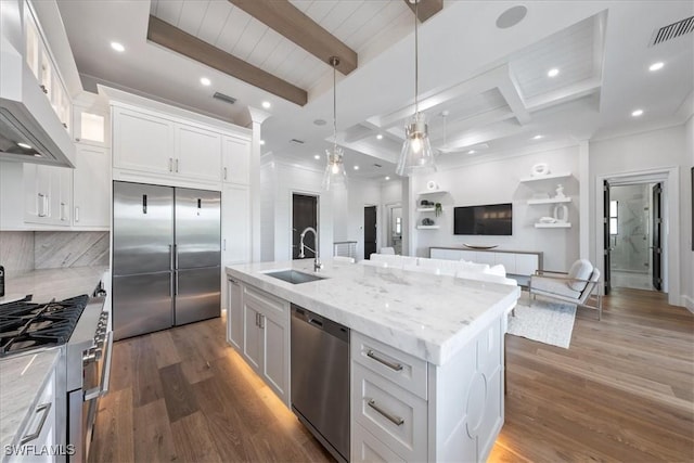 kitchen with appliances with stainless steel finishes, wall chimney range hood, an island with sink, sink, and beam ceiling