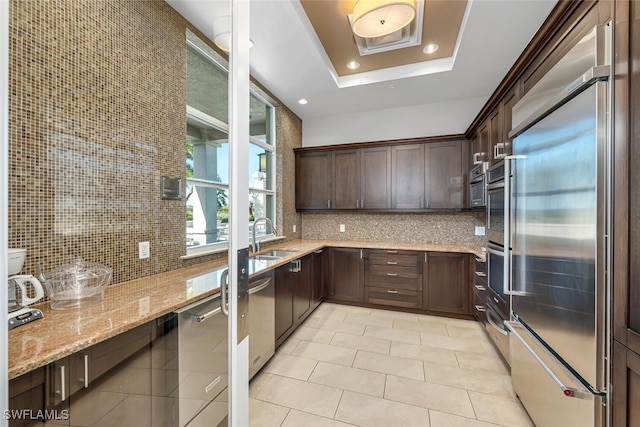 kitchen with sink, backsplash, stainless steel appliances, light stone countertops, and a raised ceiling