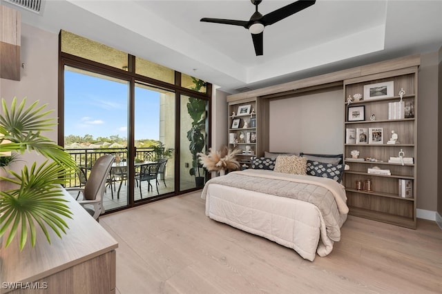 bedroom with access to outside, light wood-type flooring, a tray ceiling, expansive windows, and ceiling fan