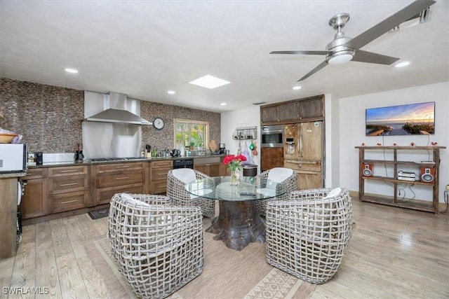 interior space featuring wall chimney range hood, light wood-type flooring, ceiling fan, and appliances with stainless steel finishes