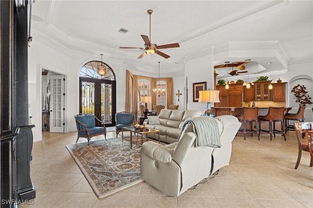 tiled living room featuring a raised ceiling, ornamental molding, ceiling fan with notable chandelier, and french doors