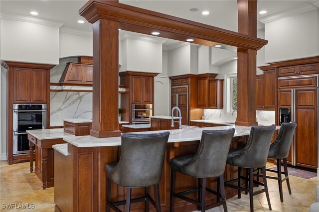 kitchen featuring a large island with sink, stainless steel double oven, crown molding, paneled built in refrigerator, and a kitchen bar