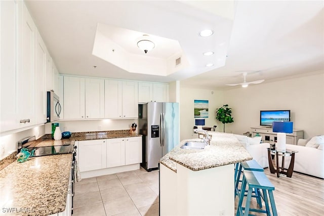 kitchen featuring a raised ceiling, sink, appliances with stainless steel finishes, white cabinetry, and a center island with sink