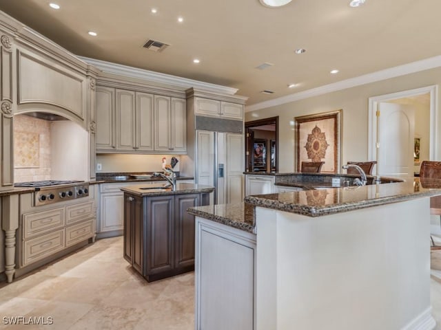 kitchen featuring stainless steel gas stovetop, an island with sink, dark stone counters, paneled built in refrigerator, and cream cabinets