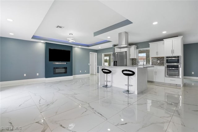 kitchen with island range hood, appliances with stainless steel finishes, a tray ceiling, a kitchen island, and white cabinets