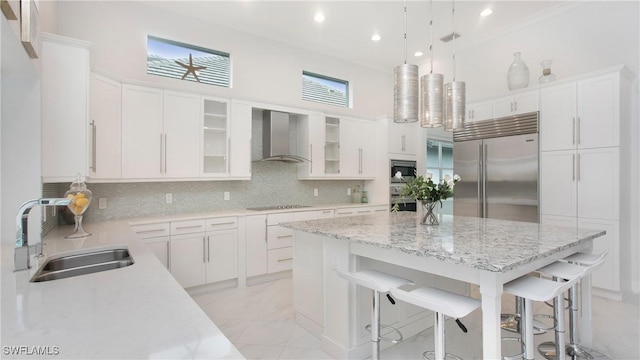 kitchen featuring wall chimney range hood, sink, built in appliances, white cabinets, and decorative light fixtures