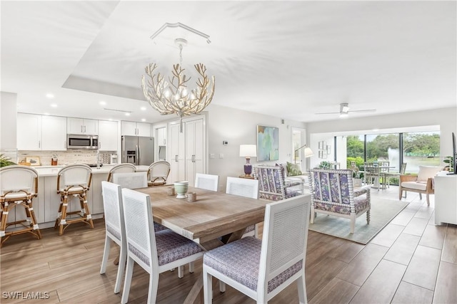 dining area with ceiling fan with notable chandelier, a raised ceiling, and light wood-type flooring