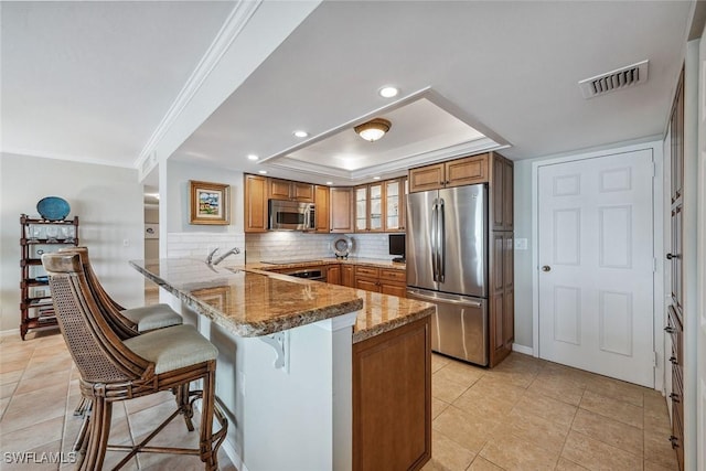 kitchen featuring a kitchen bar, a tray ceiling, kitchen peninsula, stainless steel appliances, and light stone countertops